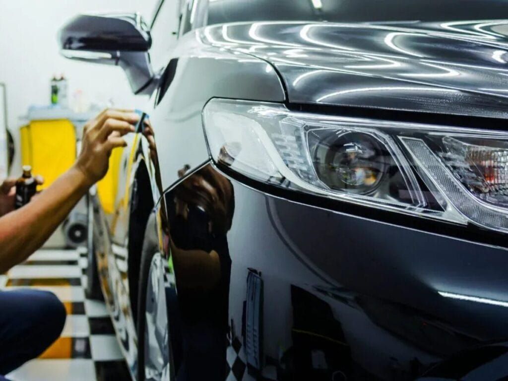 Person applying protective film to a black car in a detailing shop.