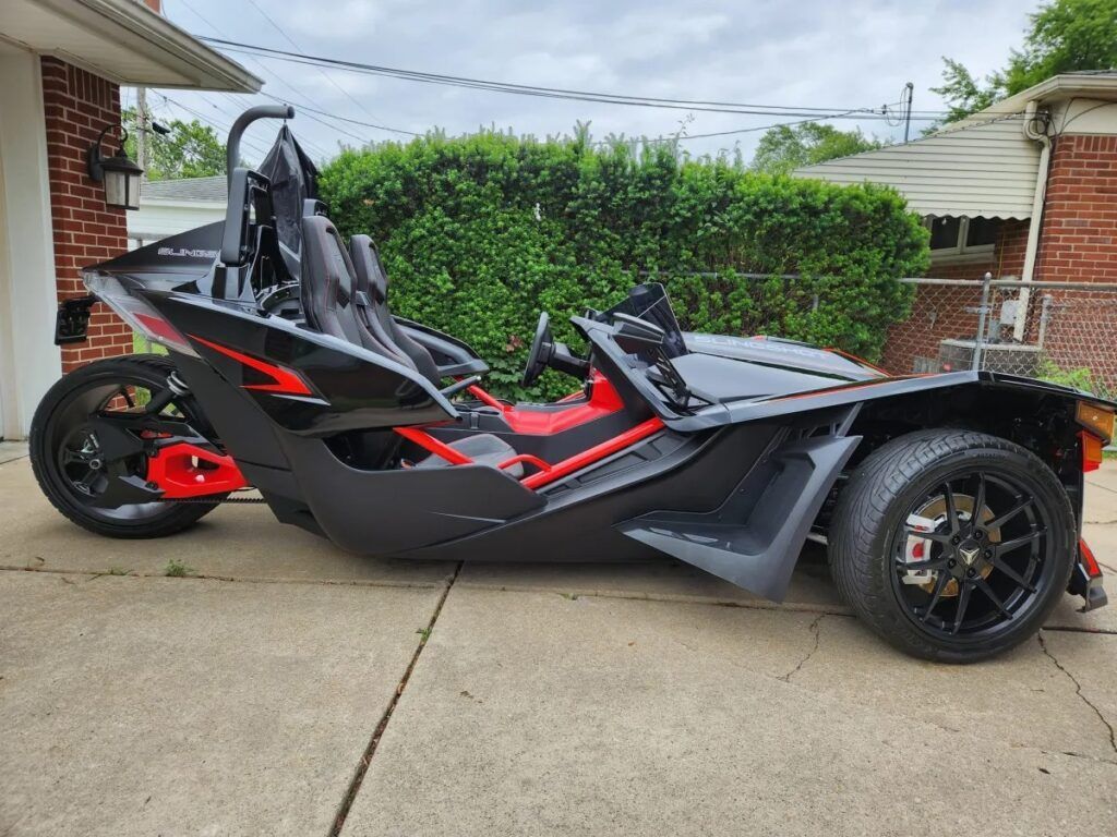 Black and red Polaris Slingshot trike parked on a driveway, with a brick house and green bushes in the background.