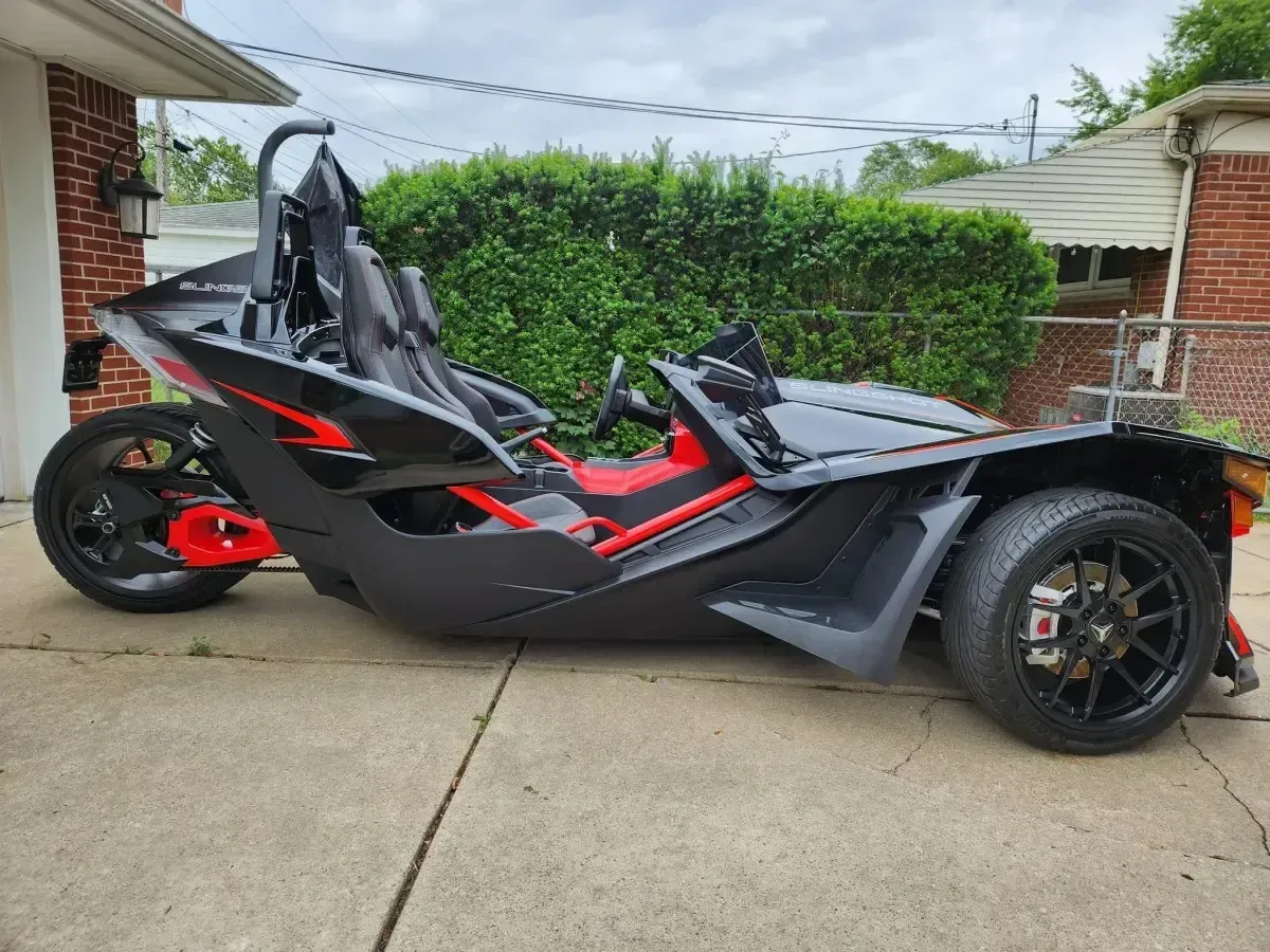 Black and red three-wheeled Polaris Slingshot roadster parked on concrete in front of a brick house and green hedges.