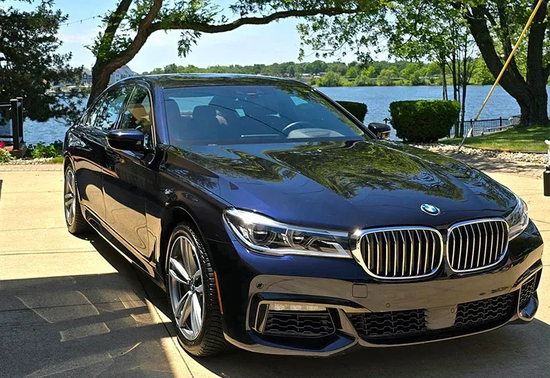 Dark blue BMW sedan parked near a lake, under a tree on a sunny day.