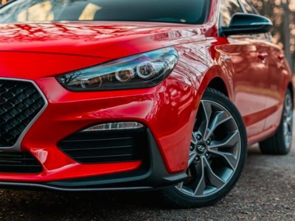 Red Hyundai car, front view; dark gray wheels, forest background.