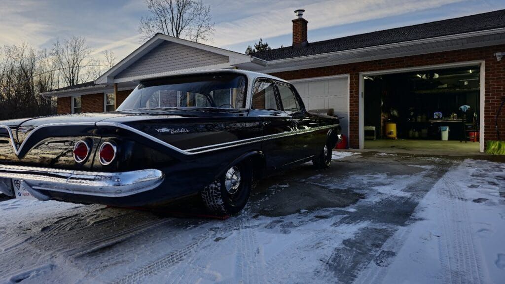 Black classic car parked on snowy driveway in front of a brick house with an open garage.