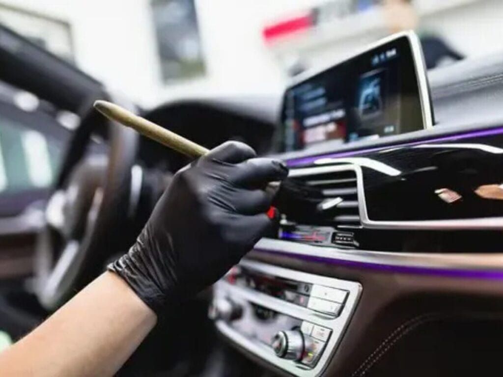 A person in a black glove cleaning a car's dashboard vents with a small brush.