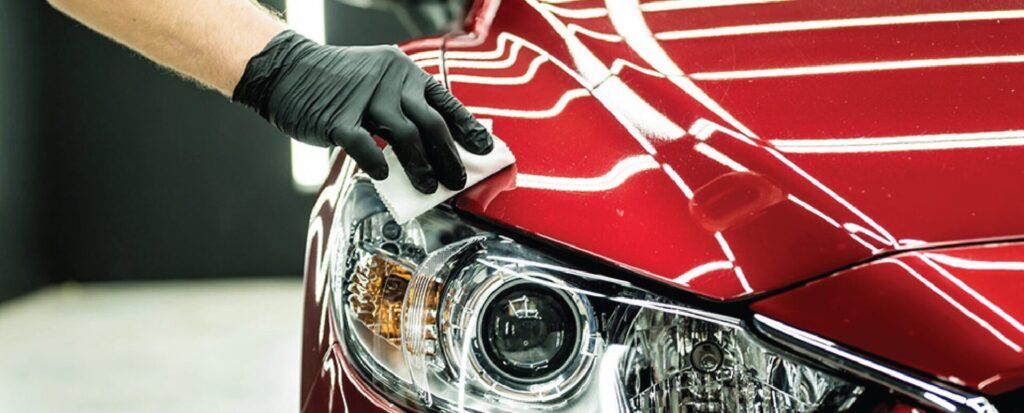 Person wearing a black glove is wiping a red car's headlight with a white cloth.