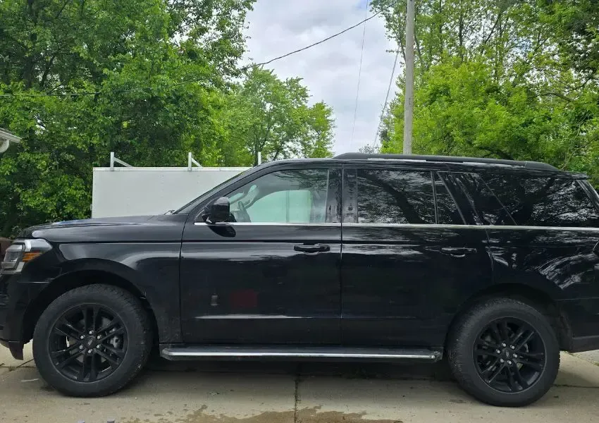 Black Ford Expedition SUV parked on a driveway, black wheels, in front of a white structure and green trees.