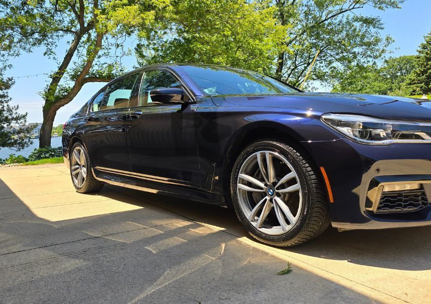 Blue BMW sedan parked on a concrete driveway with a water view on a sunny day.