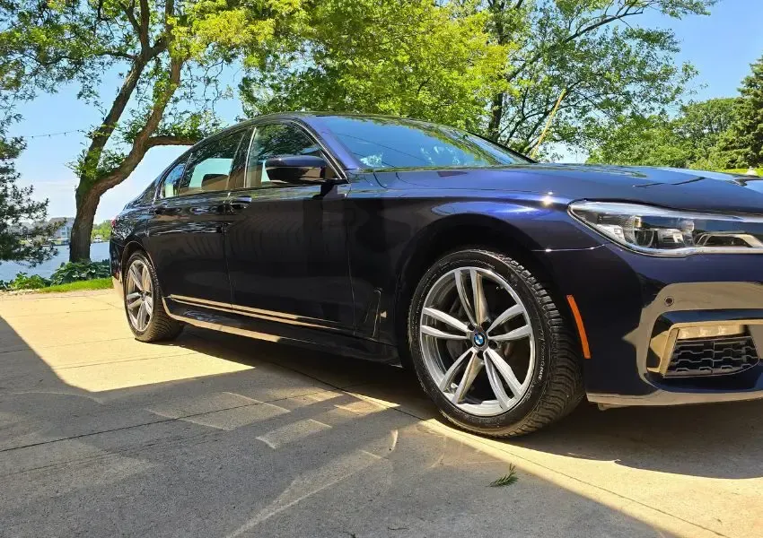 Blue BMW 7 Series sedan parked on a driveway. Daytime shot with a waterfront backdrop.