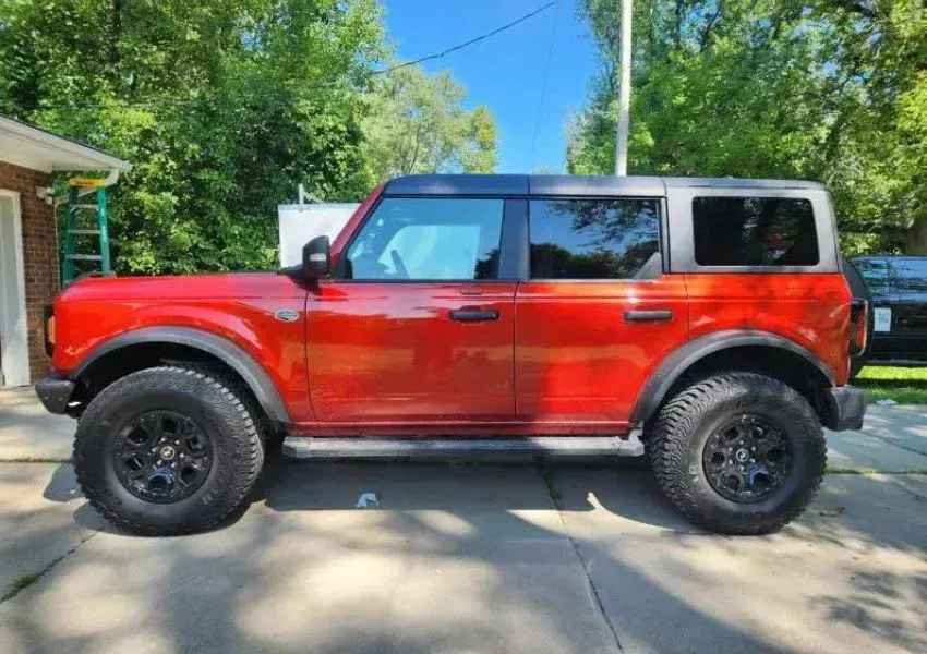 Red Ford Bronco SUV parked on a driveway, black wheels, blue sky, green trees in background.