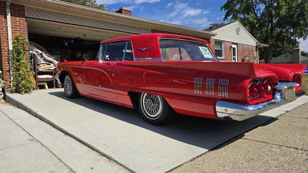 Red vintage Ford Thunderbird parked in a driveway near a garage on a sunny day.