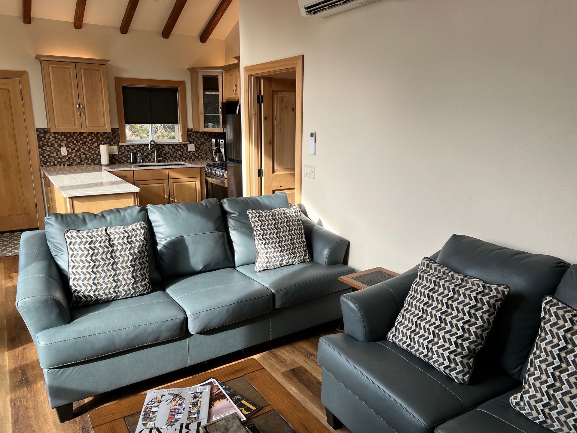 Living room with blue leather sofas, kitchen in background. Wooden beams and cabinets.