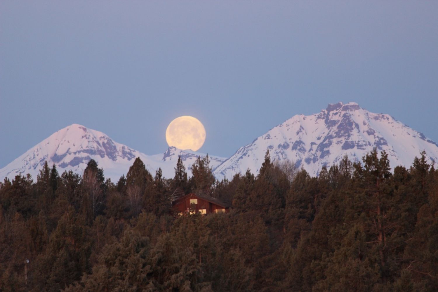Full moon rising between snow-capped mountains; a house sits among silhouetted trees.