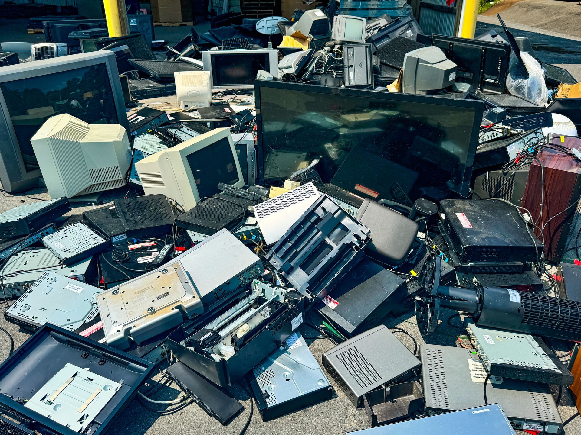 Pile of discarded electronics, including monitors, printers, and computers, on an outdoor surface.