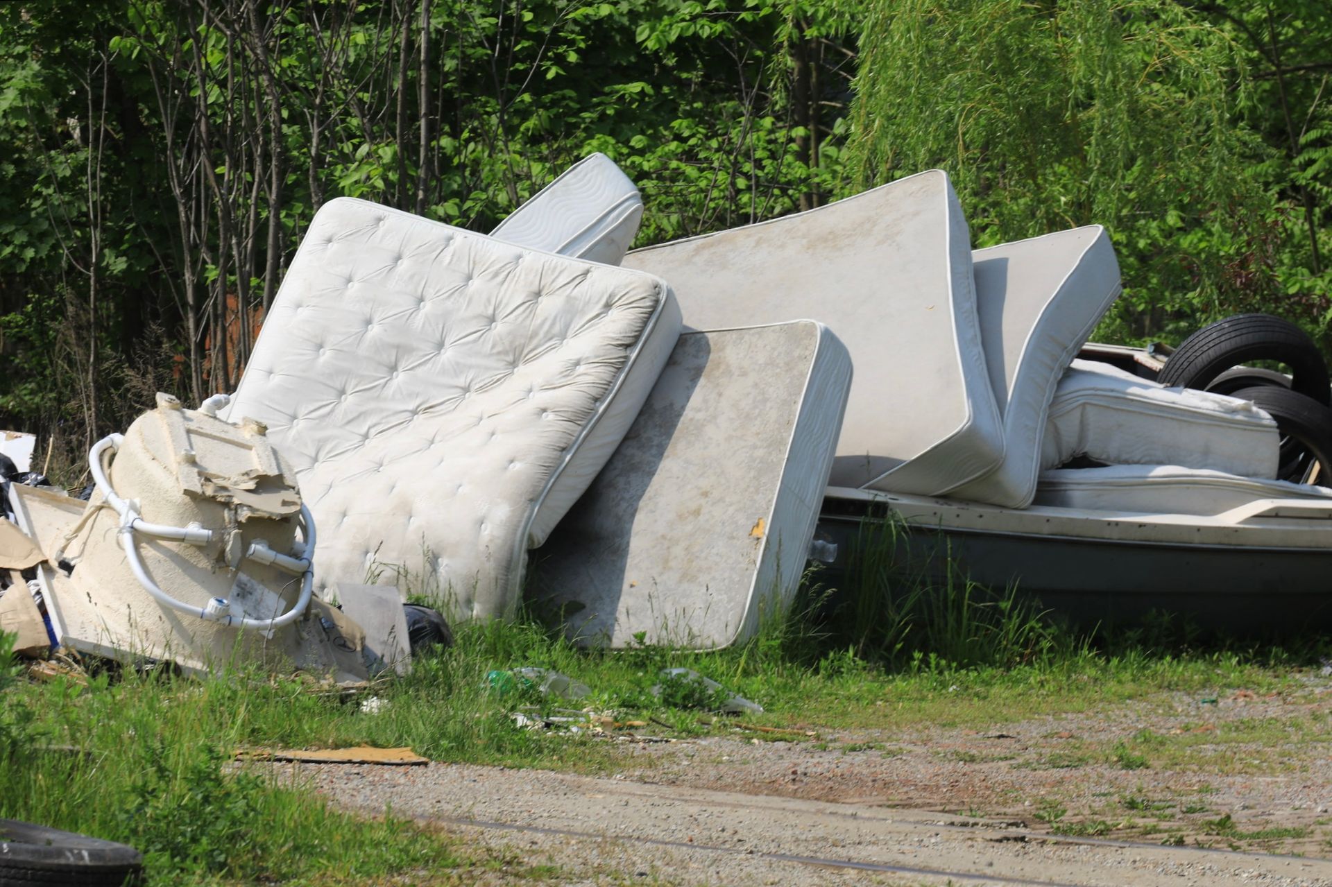 Pile of discarded mattresses and debris on the side of a gravel road near some trees.