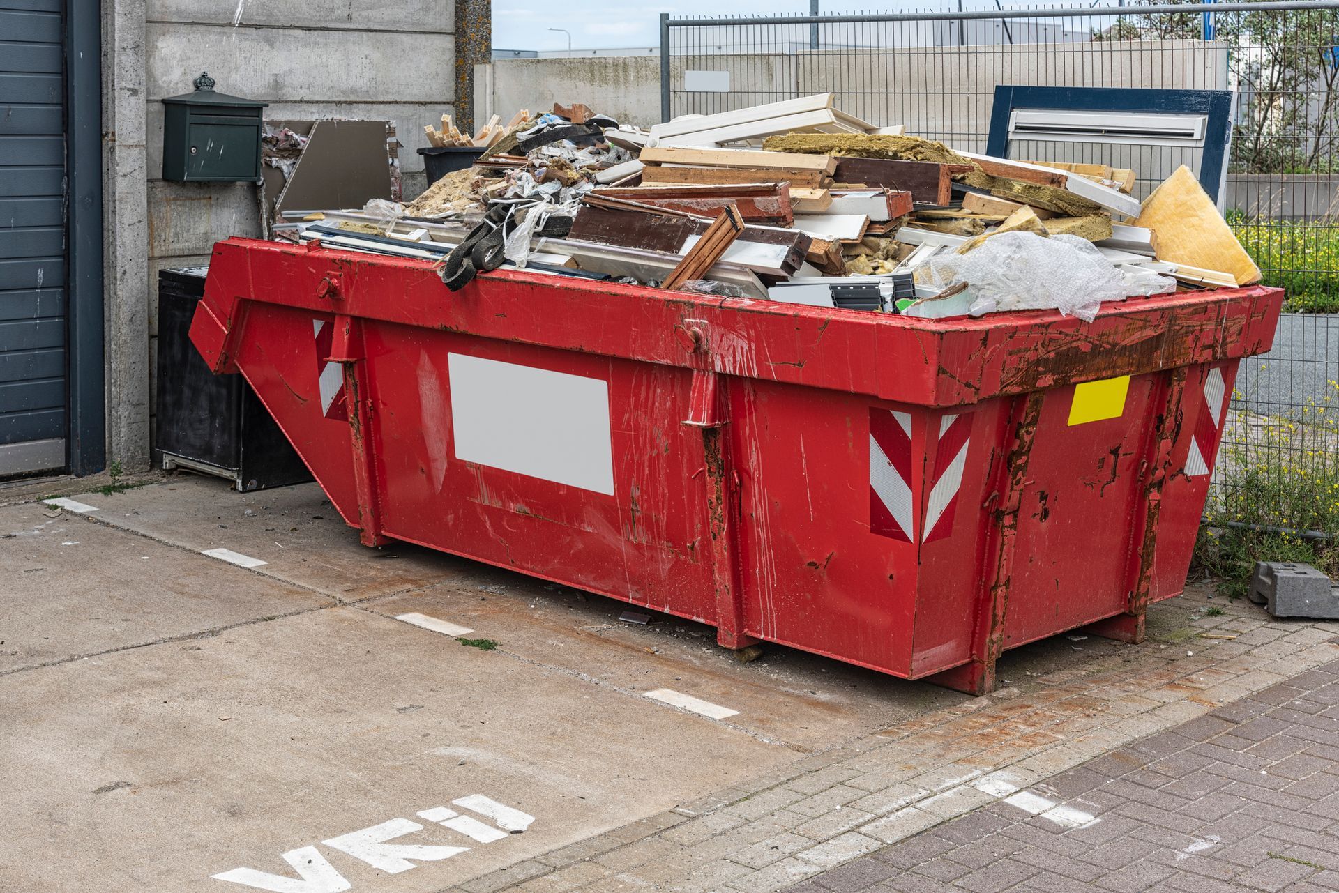 A red metal dumpster, filled with home renovation debris, stands outside a property.