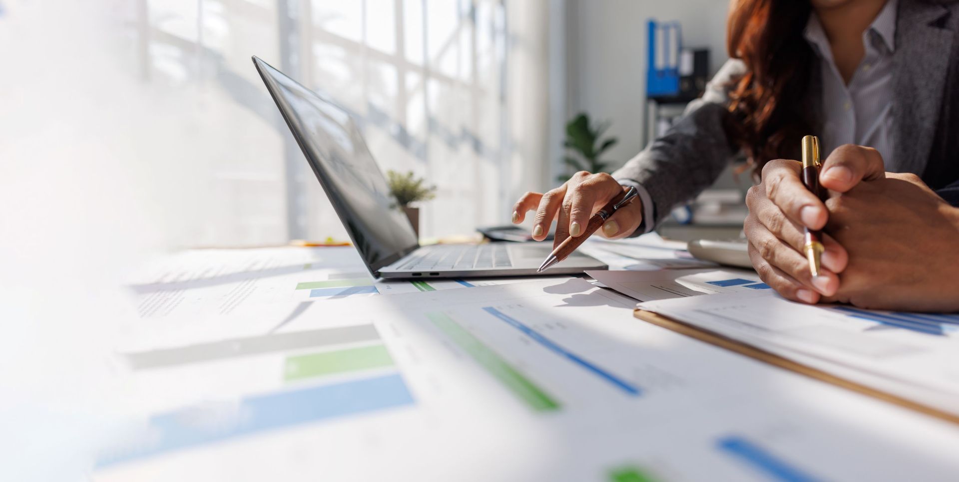 Two people review financial charts on a desk with a laptop and documents in a bright office.