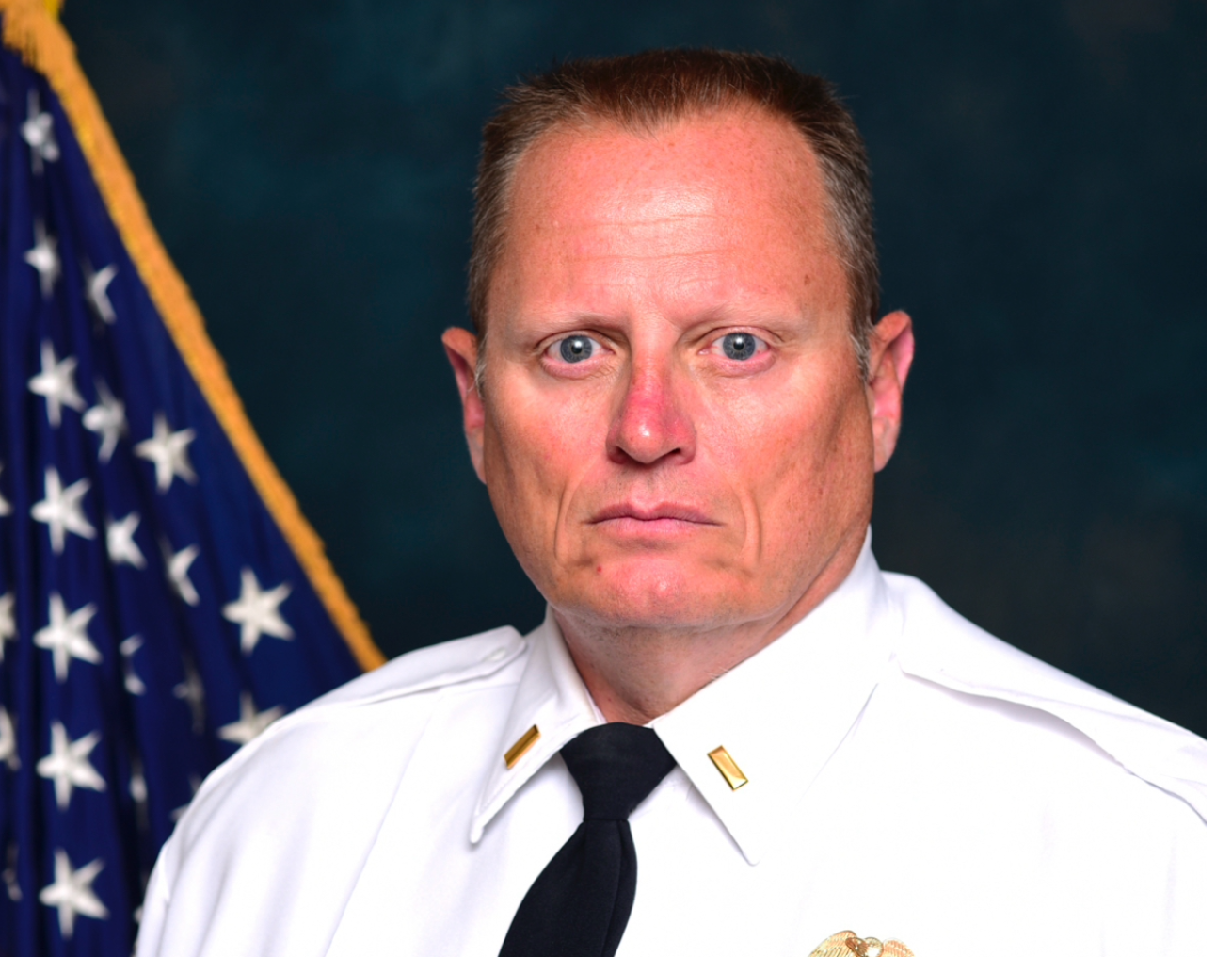 A man in a white shirt and tie stands in front of an american flag