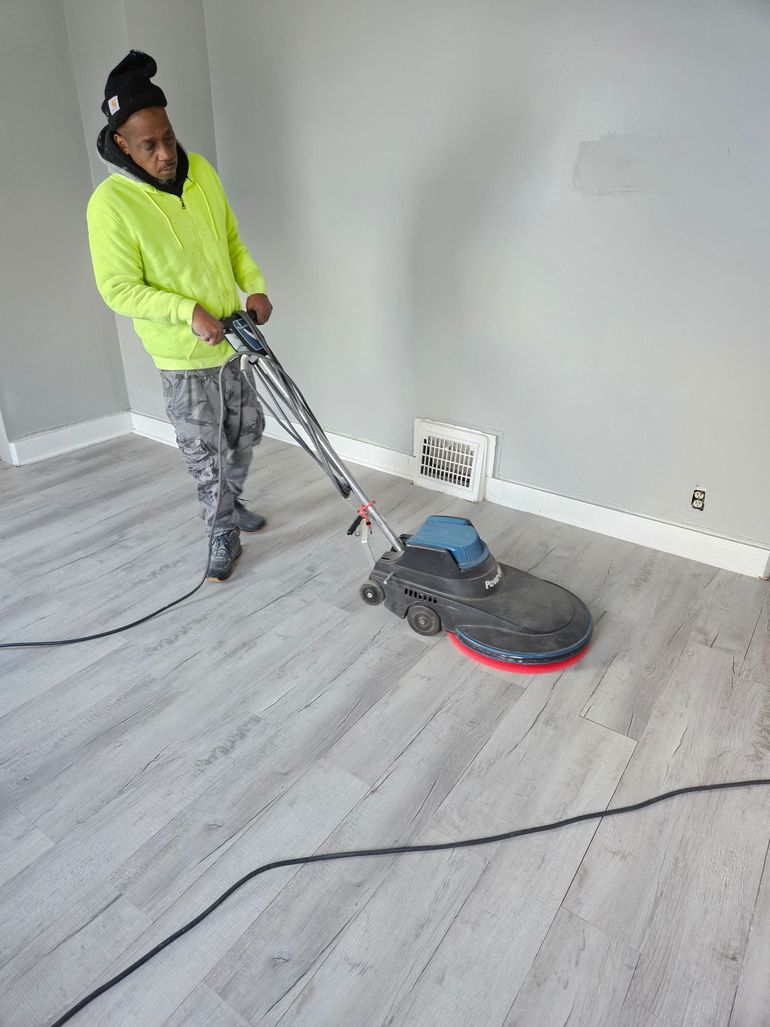 A person in a bright yellow hoodie uses a floor buffer on a light gray, wood-look floor in a room with white trim.