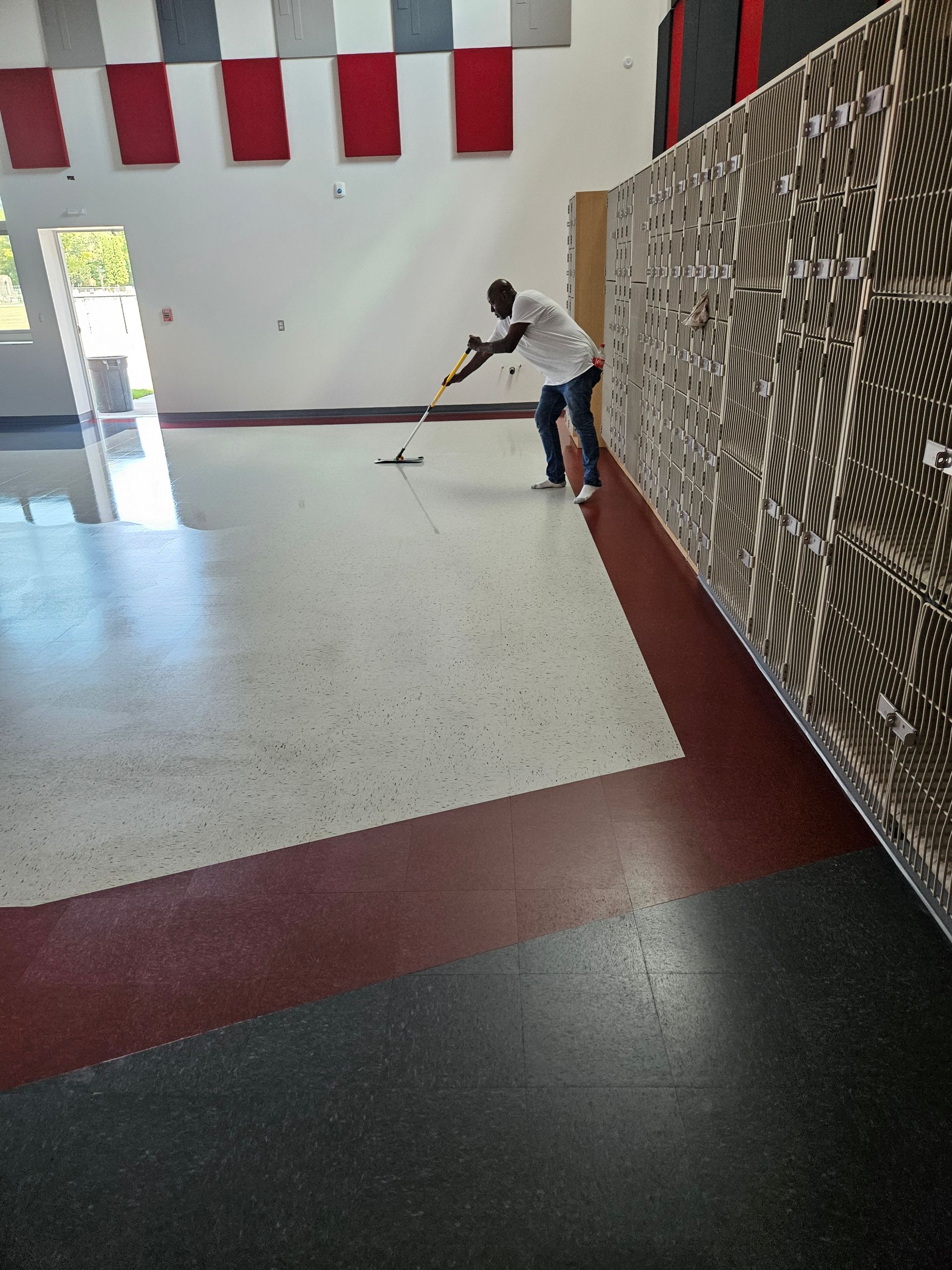 A person in a white shirt mops a speckled floor in a large room with lockers and rectangular wall accents.