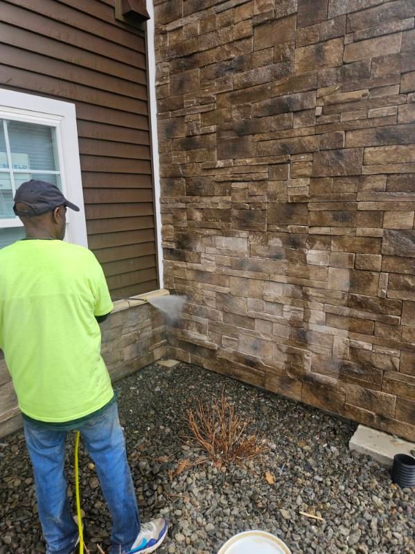 A worker in a neon green shirt uses a power washer to clean a textured stone exterior wall on a house.