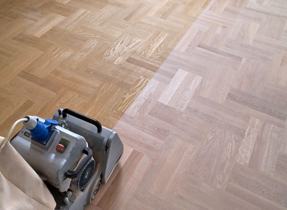 A floor sander moves across a parquet floor, showing the contrast between the worn, original finish and the sanded wood.