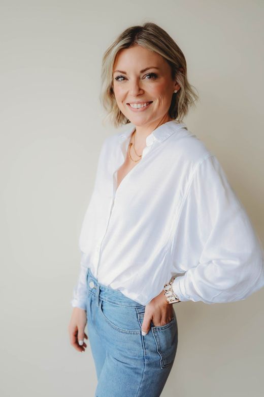 A woman wearing a white shirt and blue jeans is standing in front of a white wall.