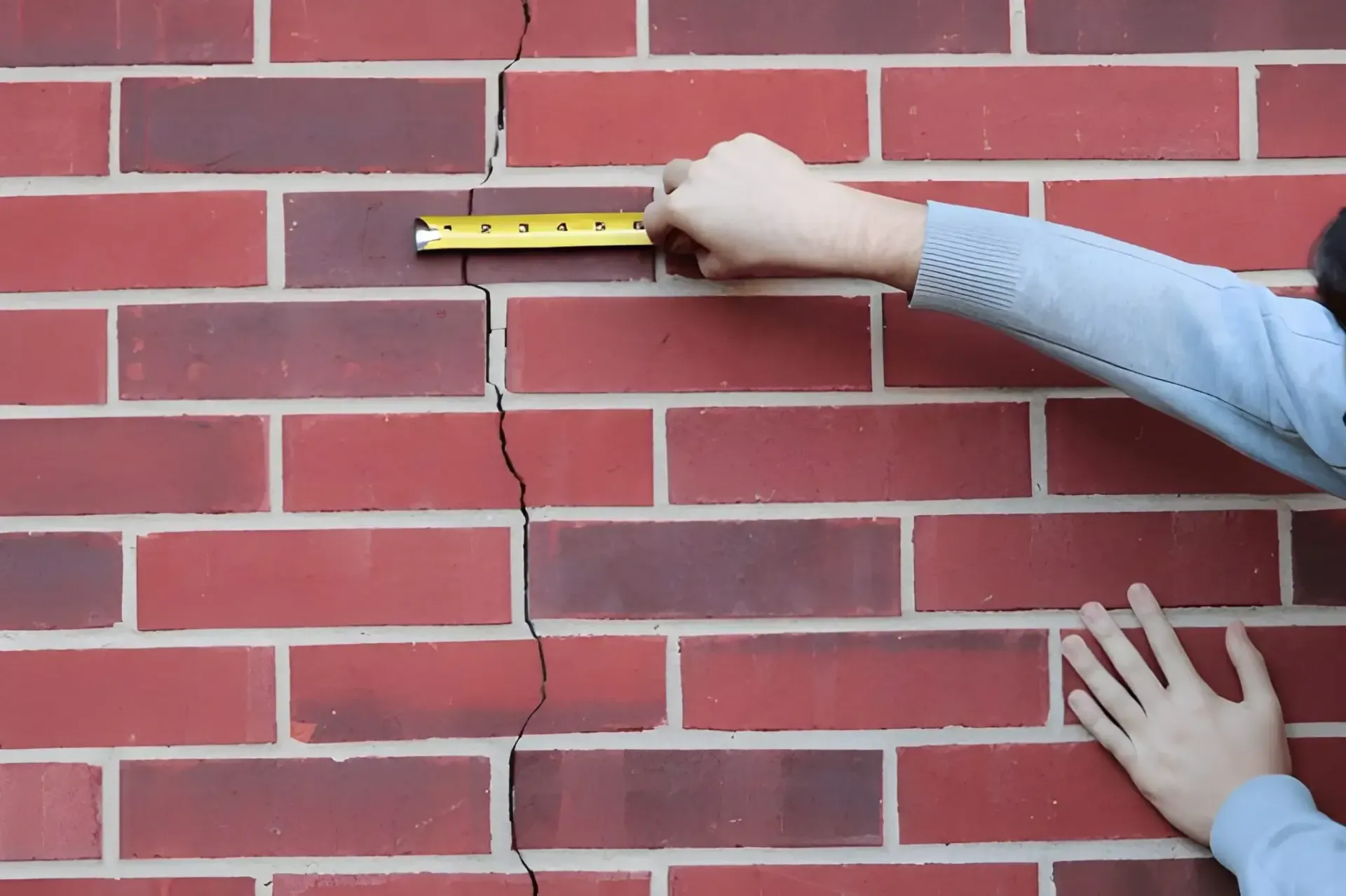 Technician measuring width of foundation crack in brick wall to determine severity and repair solutions.