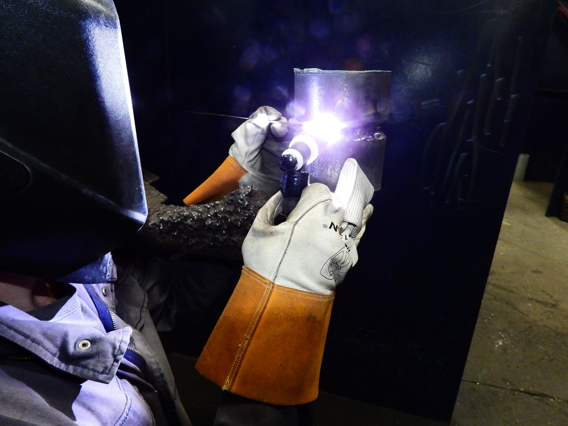 A man wearing a welding helmet and gloves is welding a pipe