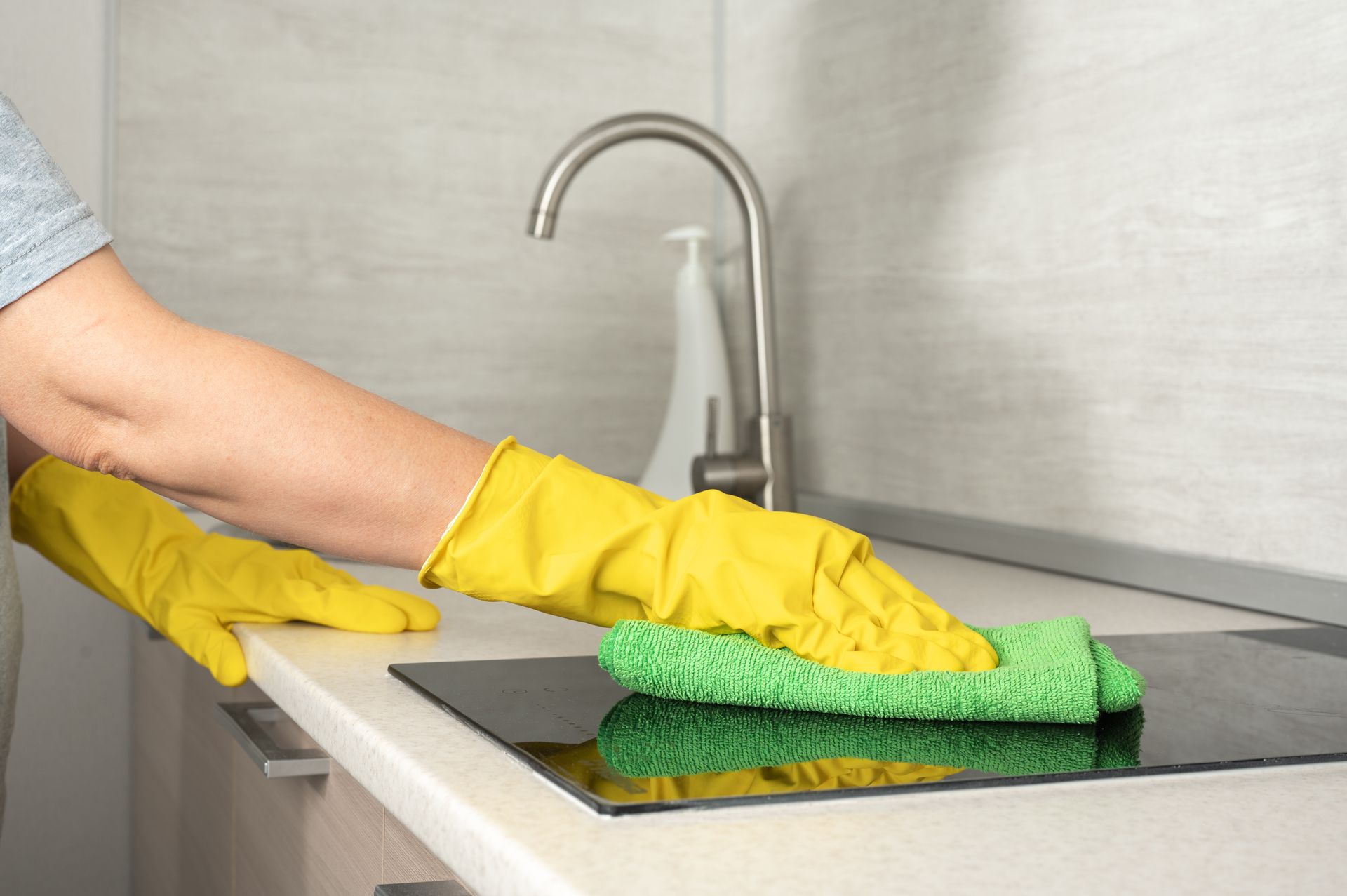 A person wearing yellow gloves is cleaning a kitchen counter with a green sponge.