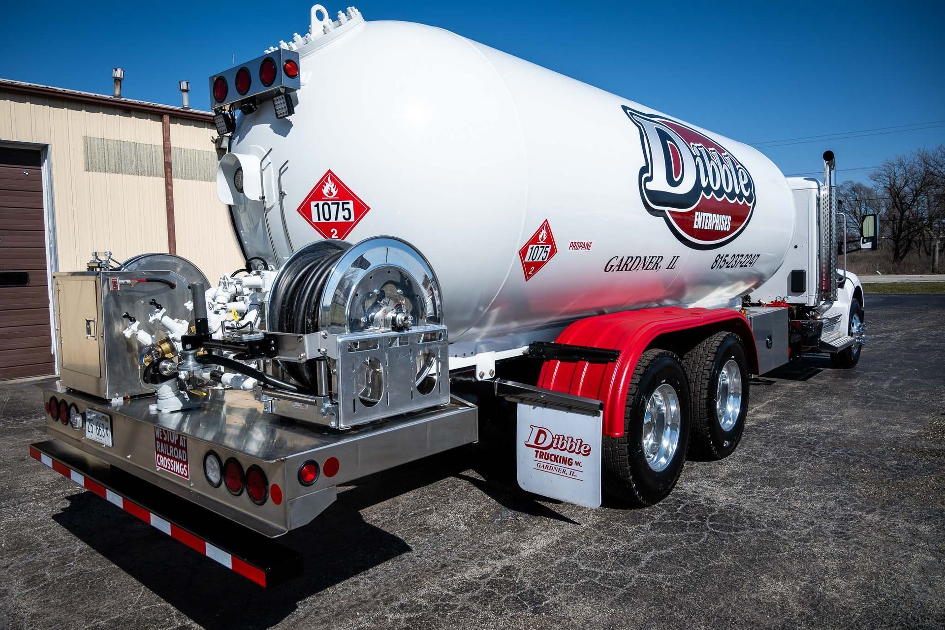 A white tanker truck with a red trailer is parked in front of a building.