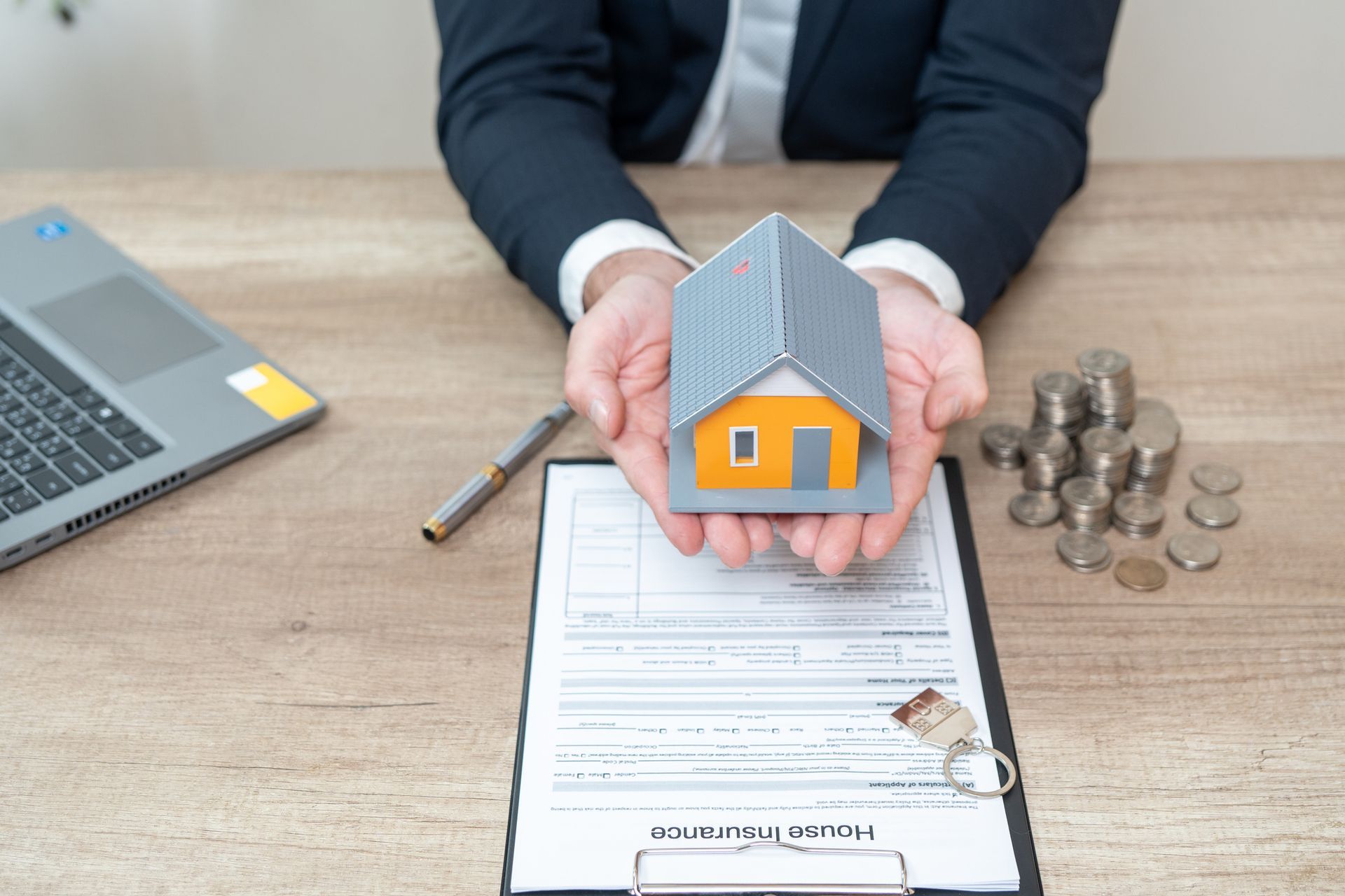 Person holding a model house, paperwork, keys, coins, and laptop on a desk.