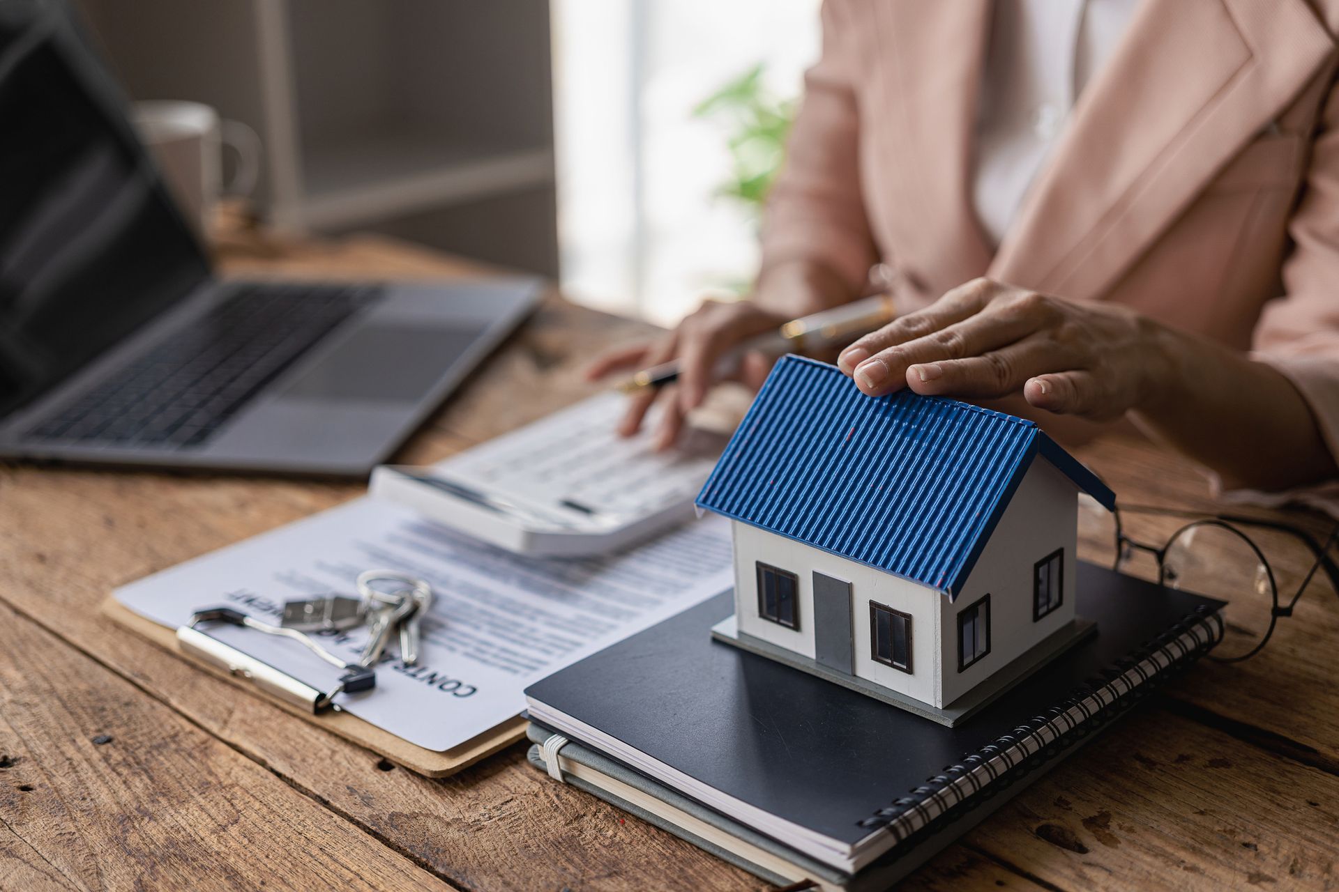 Person calculating with calculator, hands on model house, paperwork, keys, laptop on wooden desk.