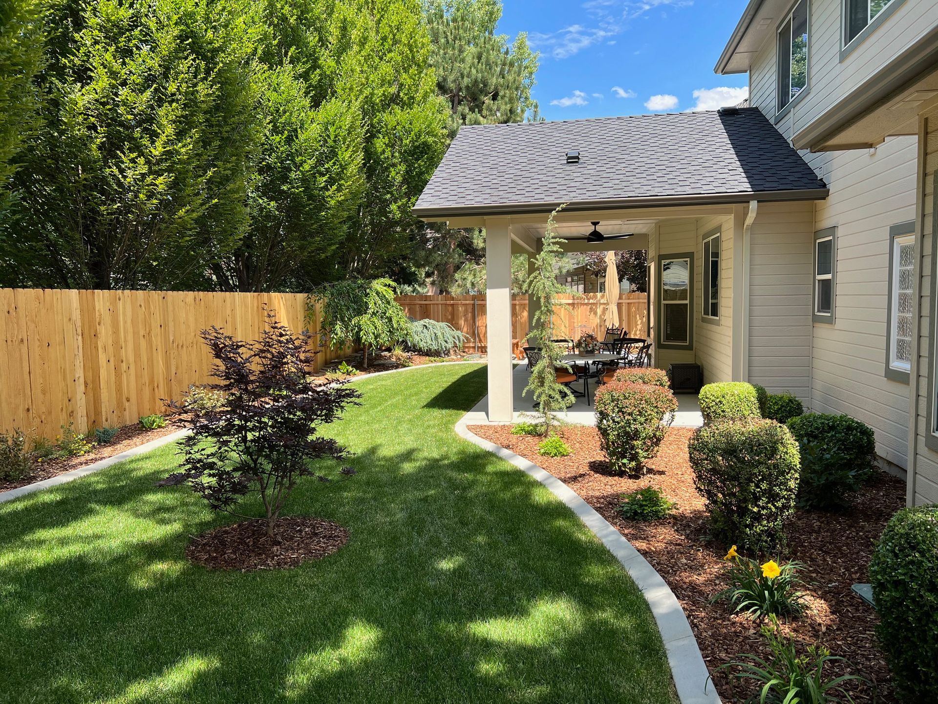 Landscaped backyard with curved stone edging, flower beds, green lawn, and a covered patio beside a beige house