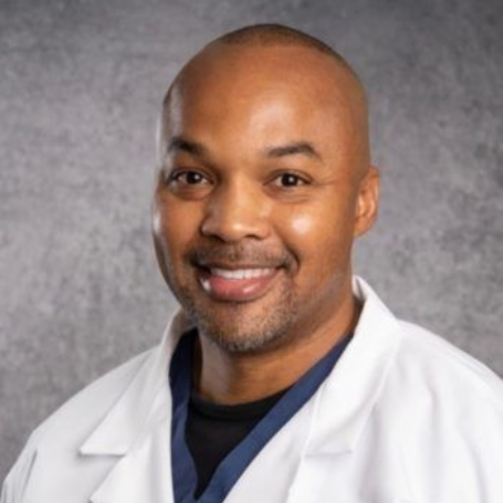 Smiling Black man in a white coat, likely a doctor, with a neutral background.