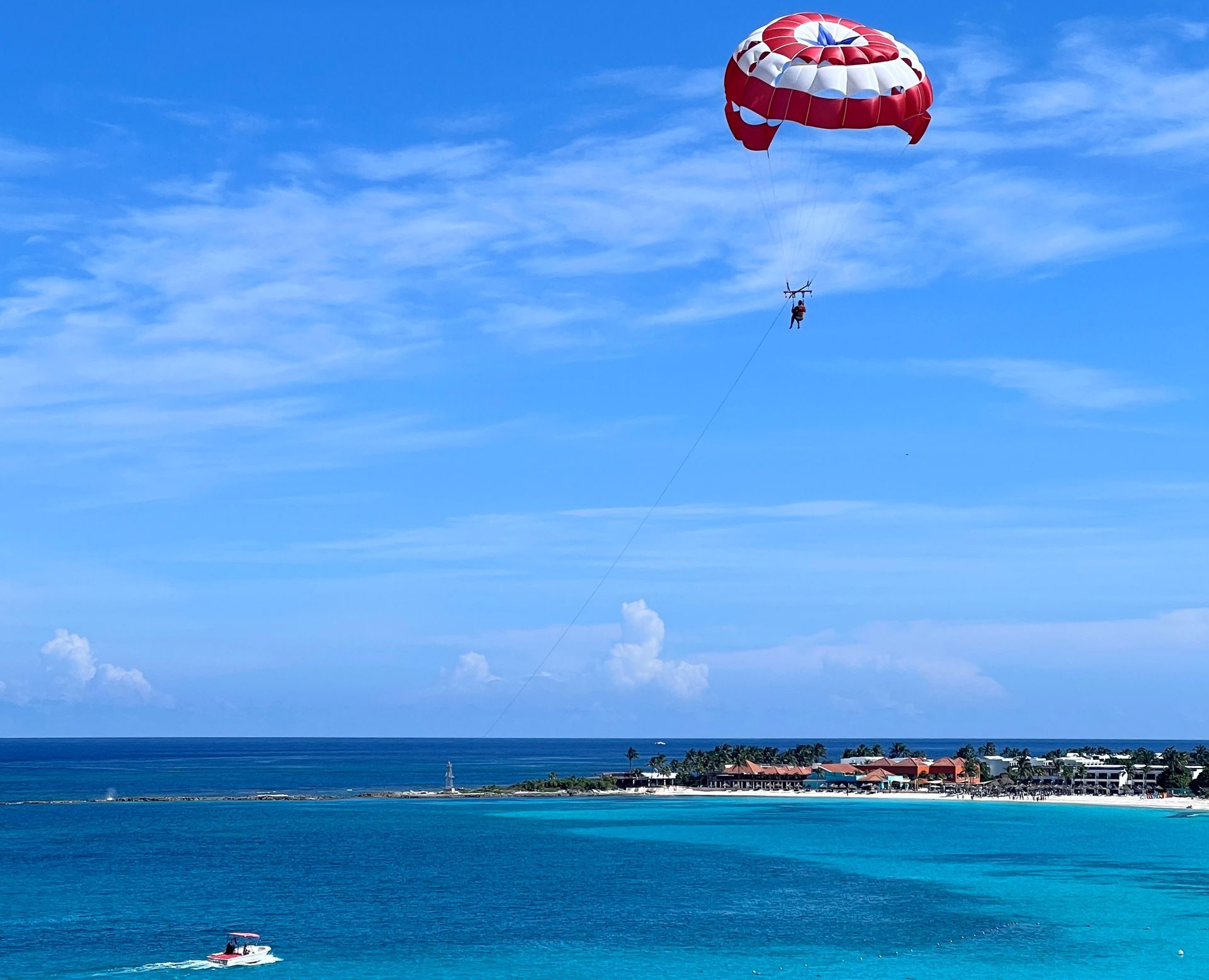 Parasailing en el cielo azul brillante sobre un océano turquesa cerca de una playa de arena blanca con edificios.