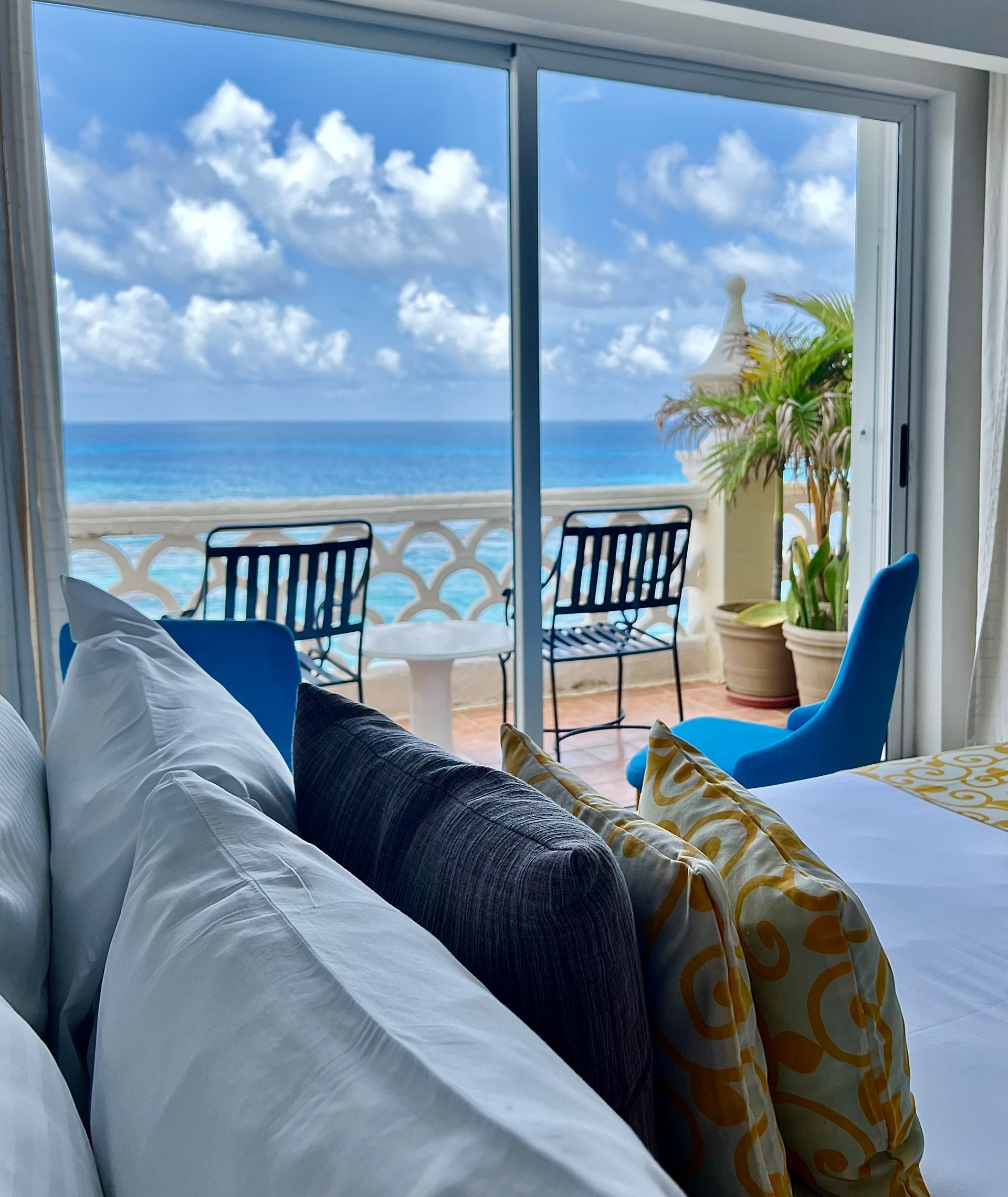Dormitorio con vista al mar, balcón con sillas, cielo azul y almohadas mullidas.