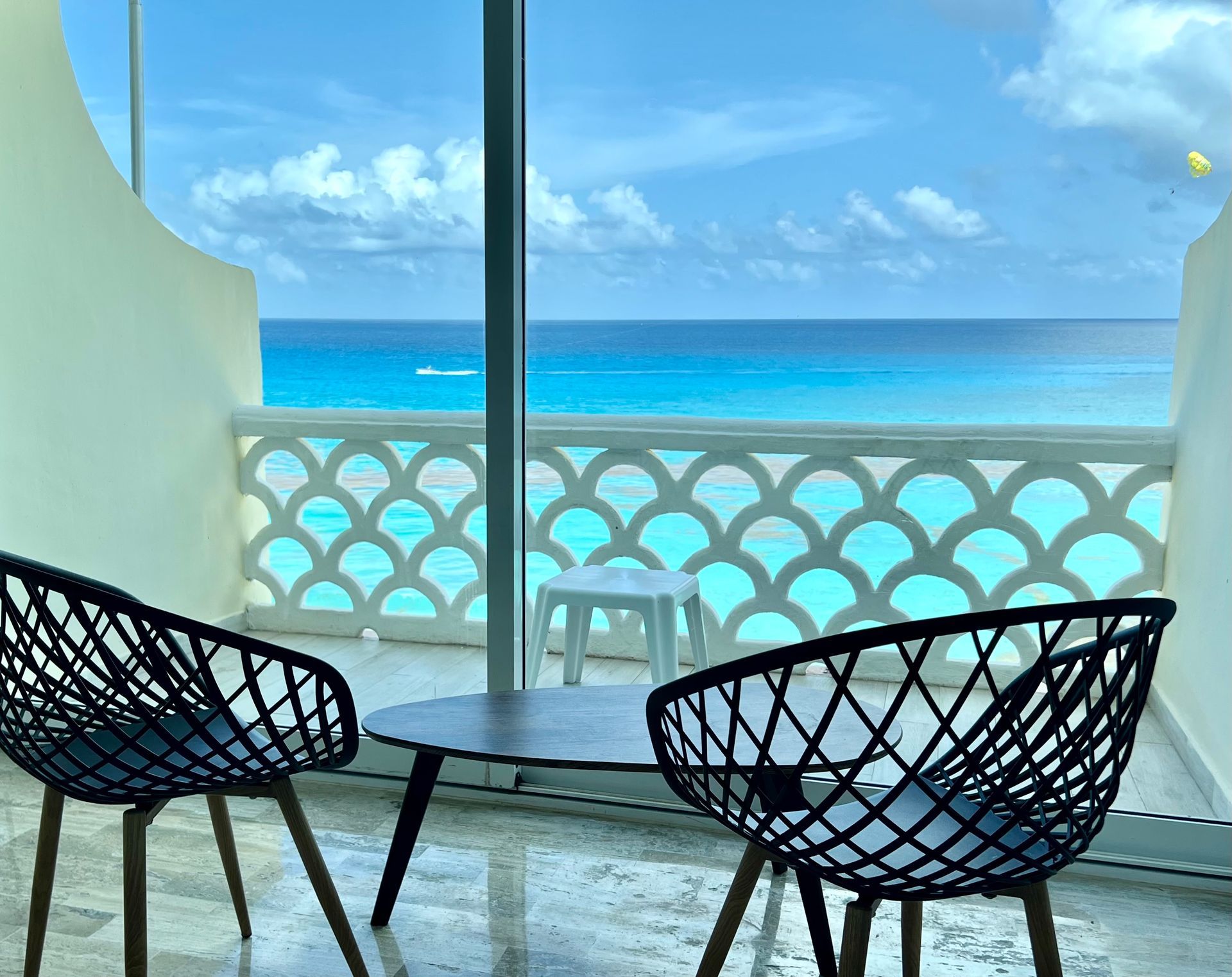 Balcón con sillas negras, mesa pequeña y vista al mar turquesa.