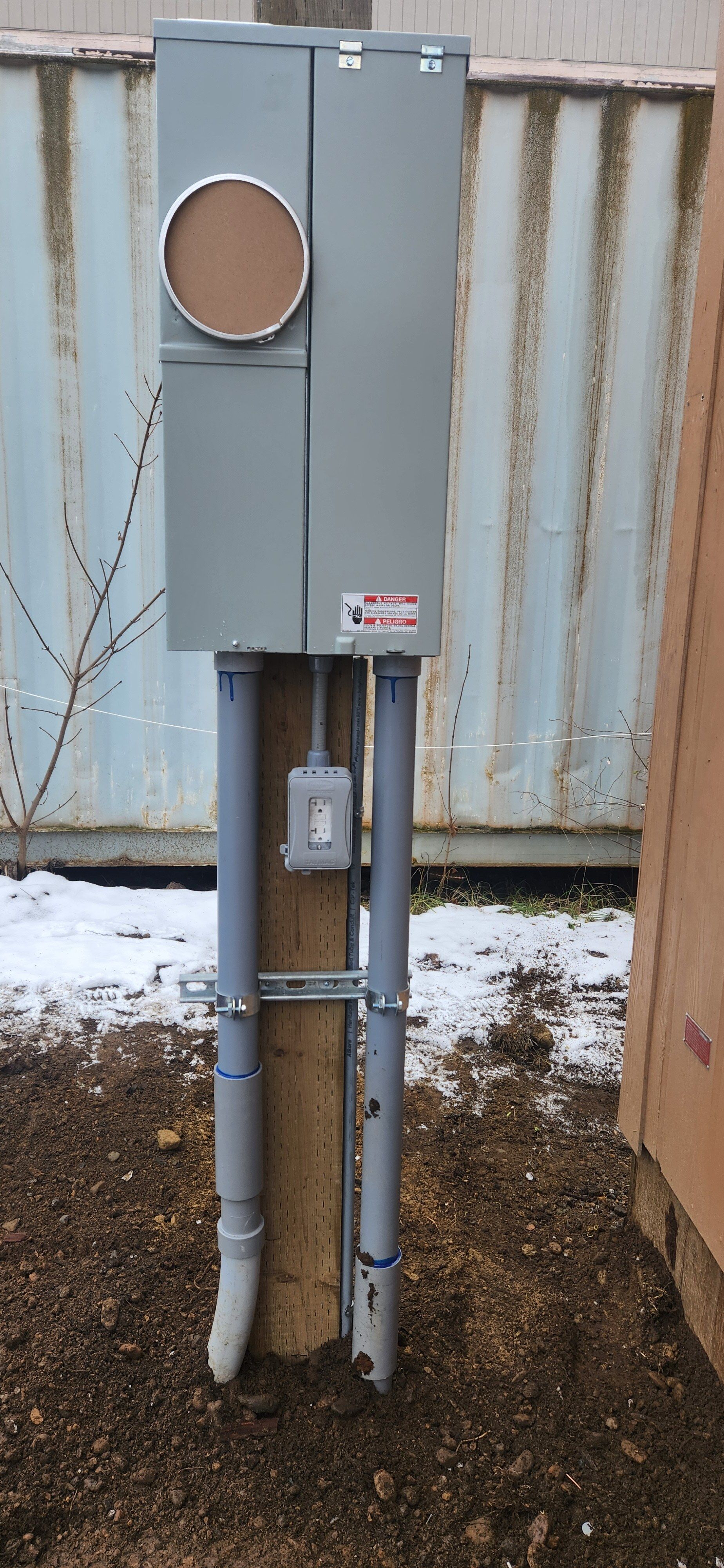 A electrical box is sitting on top of a wooden post in the dirt.