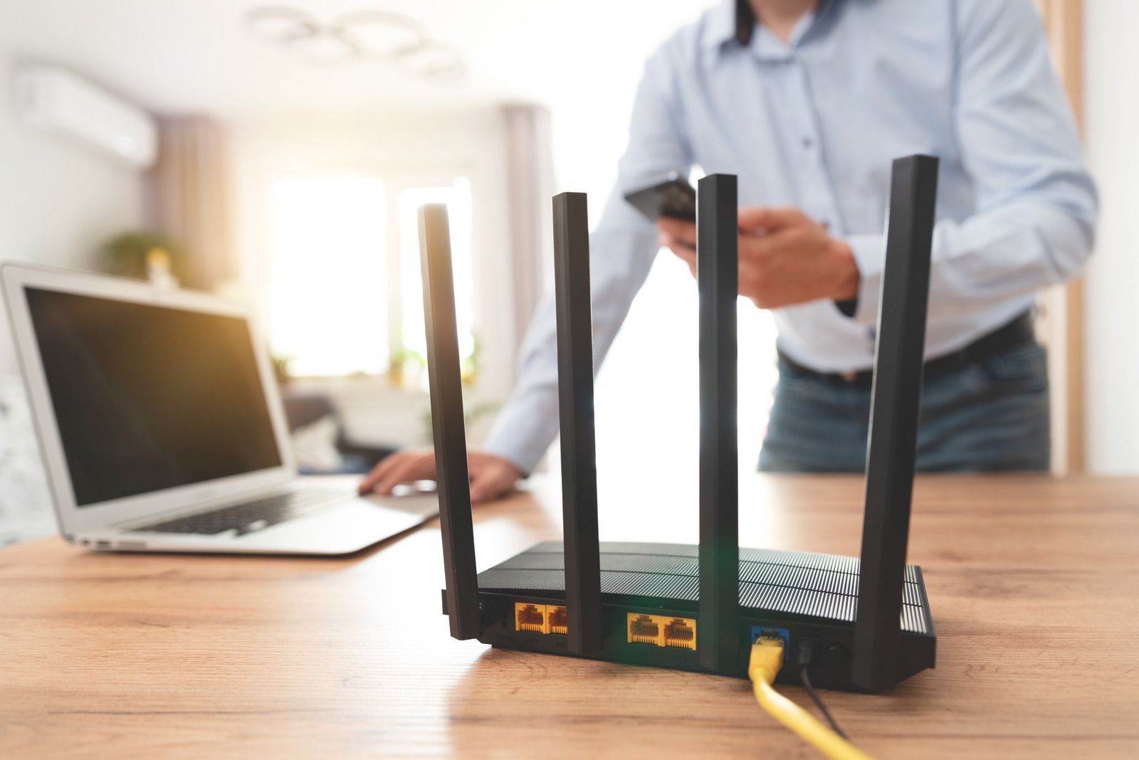 Router with antennas on a table with a laptop and a person using a phone in the background.