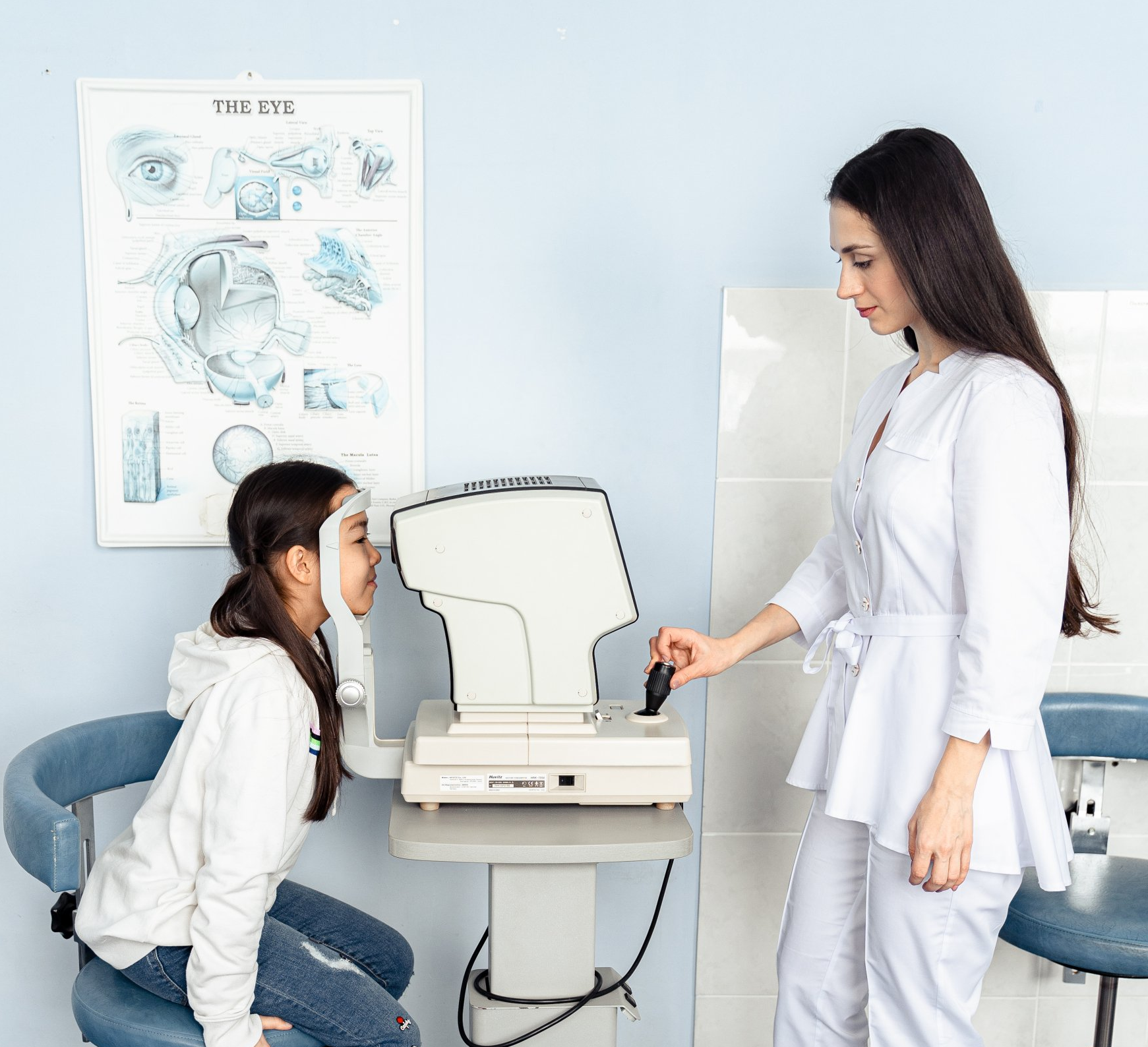 Woman examining a young person's eyes with an autorefractor in a clinic; eye chart on the wall.