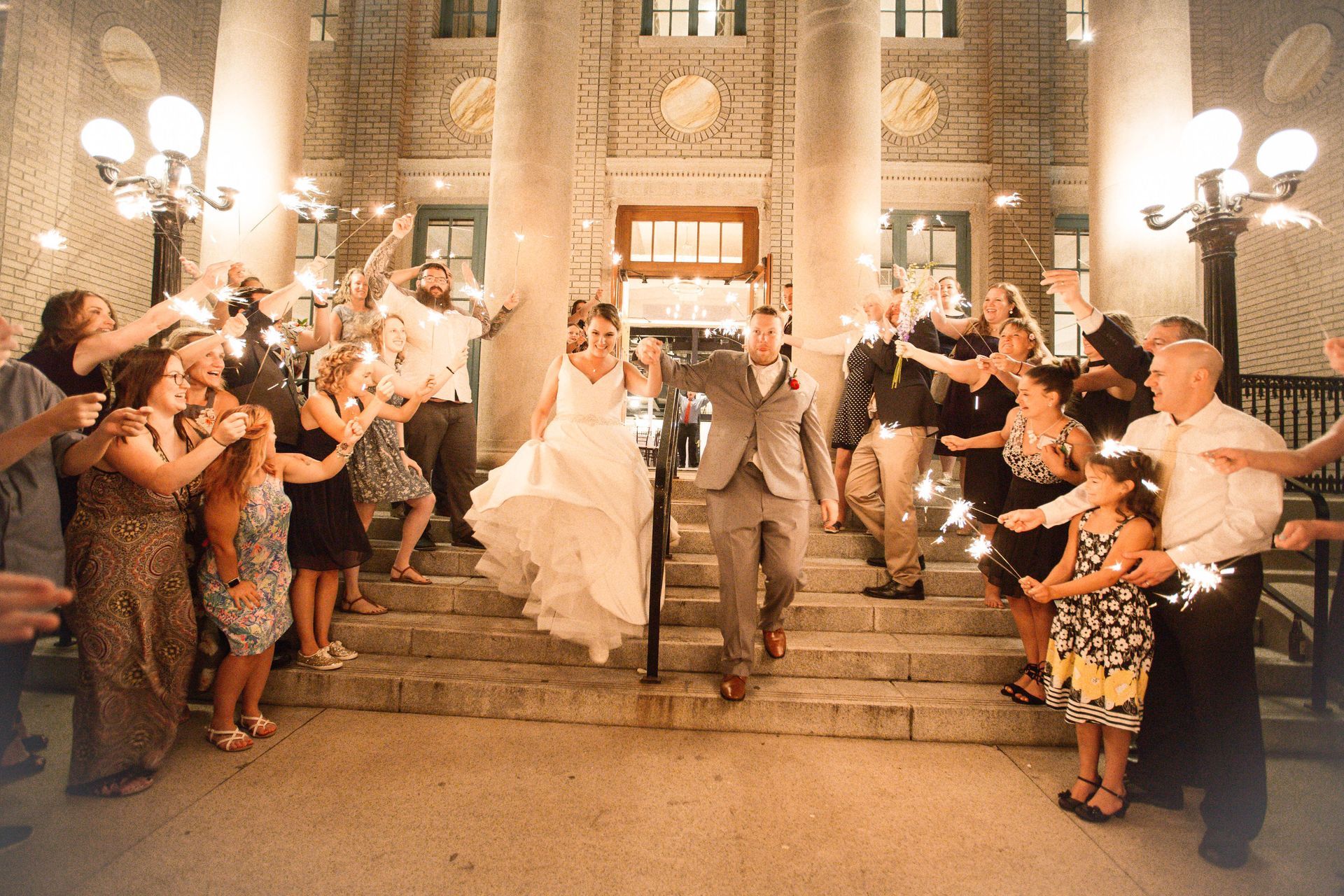 A bride and groom are walking down the stairs with their wedding party holding sparklers.