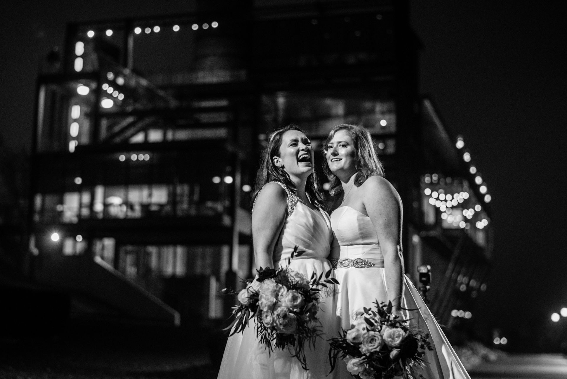 Two women in wedding dresses are standing next to each other in front of a double decker bus at night.