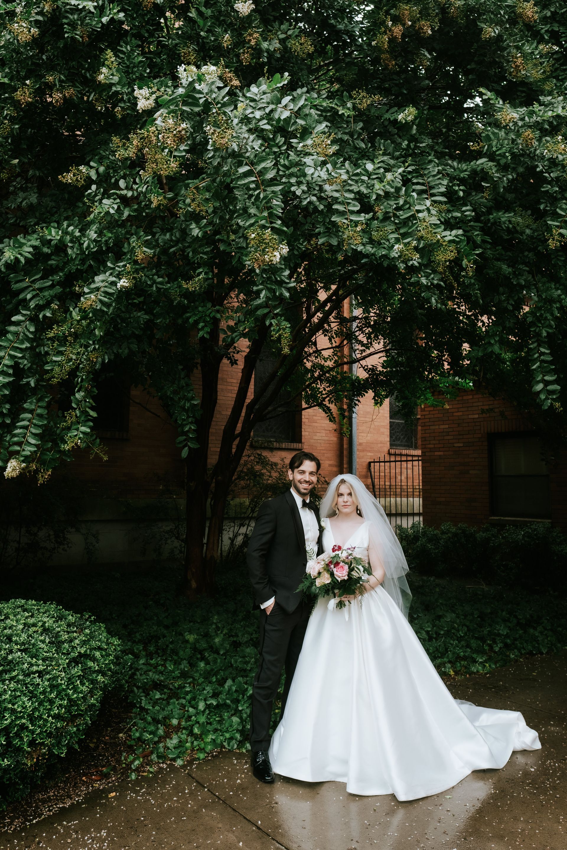 A bride and groom are standing next to each other on a sidewalk in front of a tree.