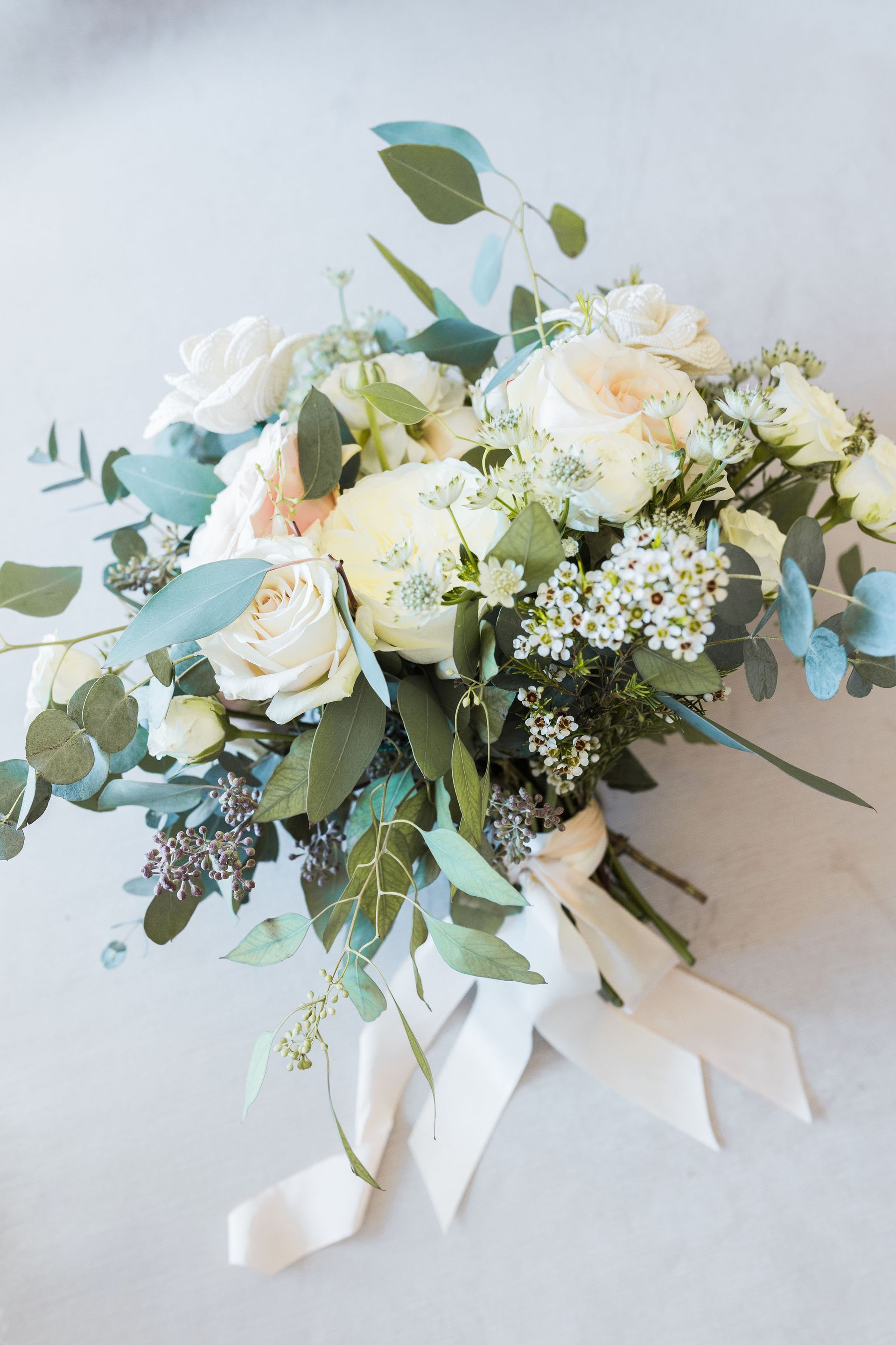 A bouquet of white flowers and greenery with a white ribbon on a table.