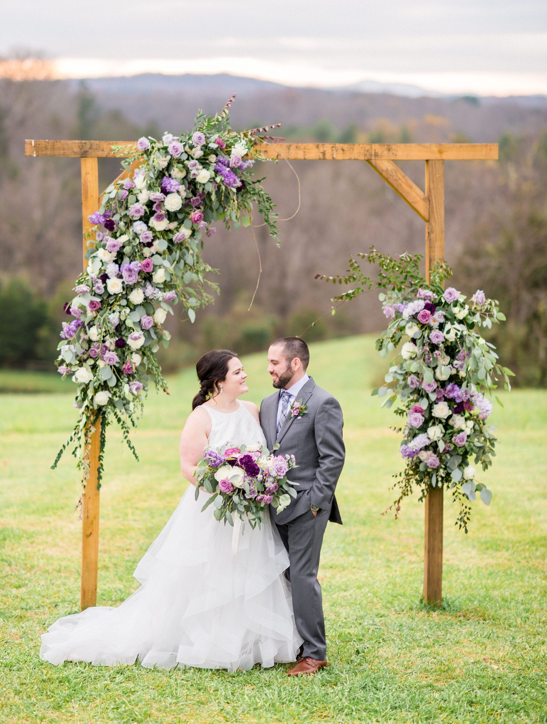 A bride and groom are standing under a wooden arch decorated with flowers.