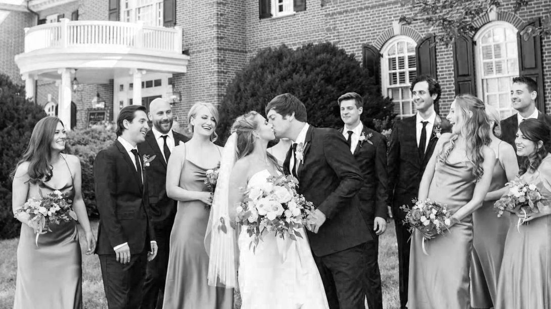 A black and white photo of a bride and groom kissing in front of their wedding party.