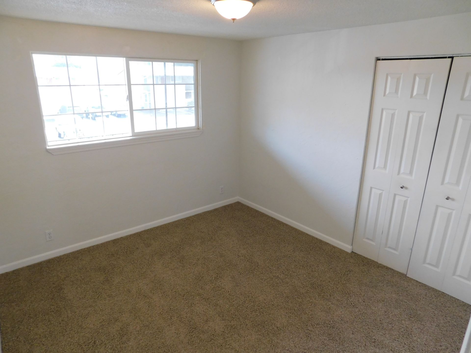 Empty bedroom with beige carpet, white walls, window, and closet doors.