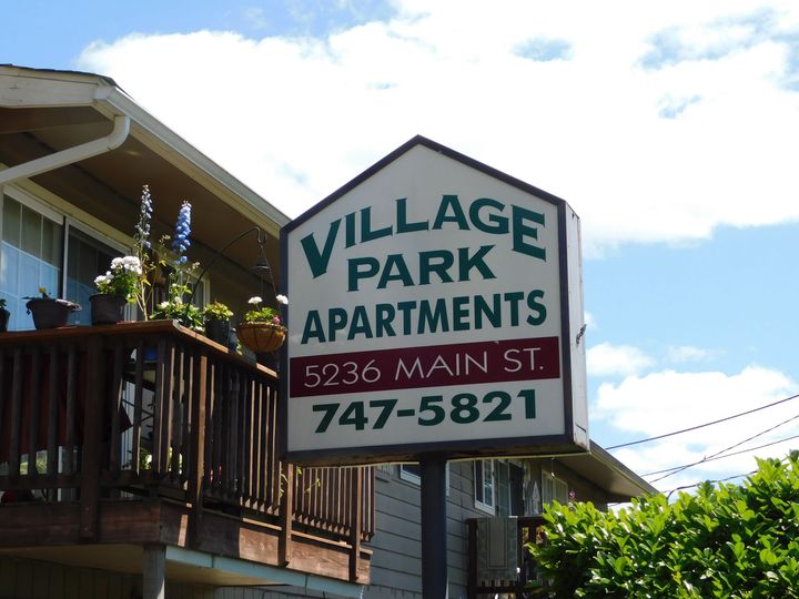 Sign for Village Park Apartments at 5936 Main St. Brown and white with green text. A building with a deck is to the left.