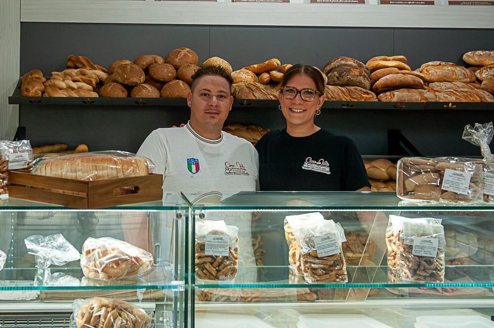 Due persone dietro il bancone di un panificio con espositori di pane.