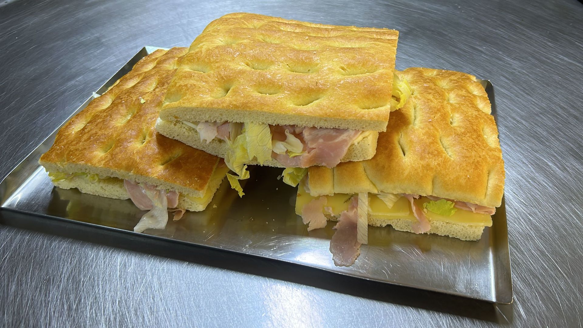 Panini su focaccia, farciti con prosciutto e lattuga, impilati su un vassoio di metallo.