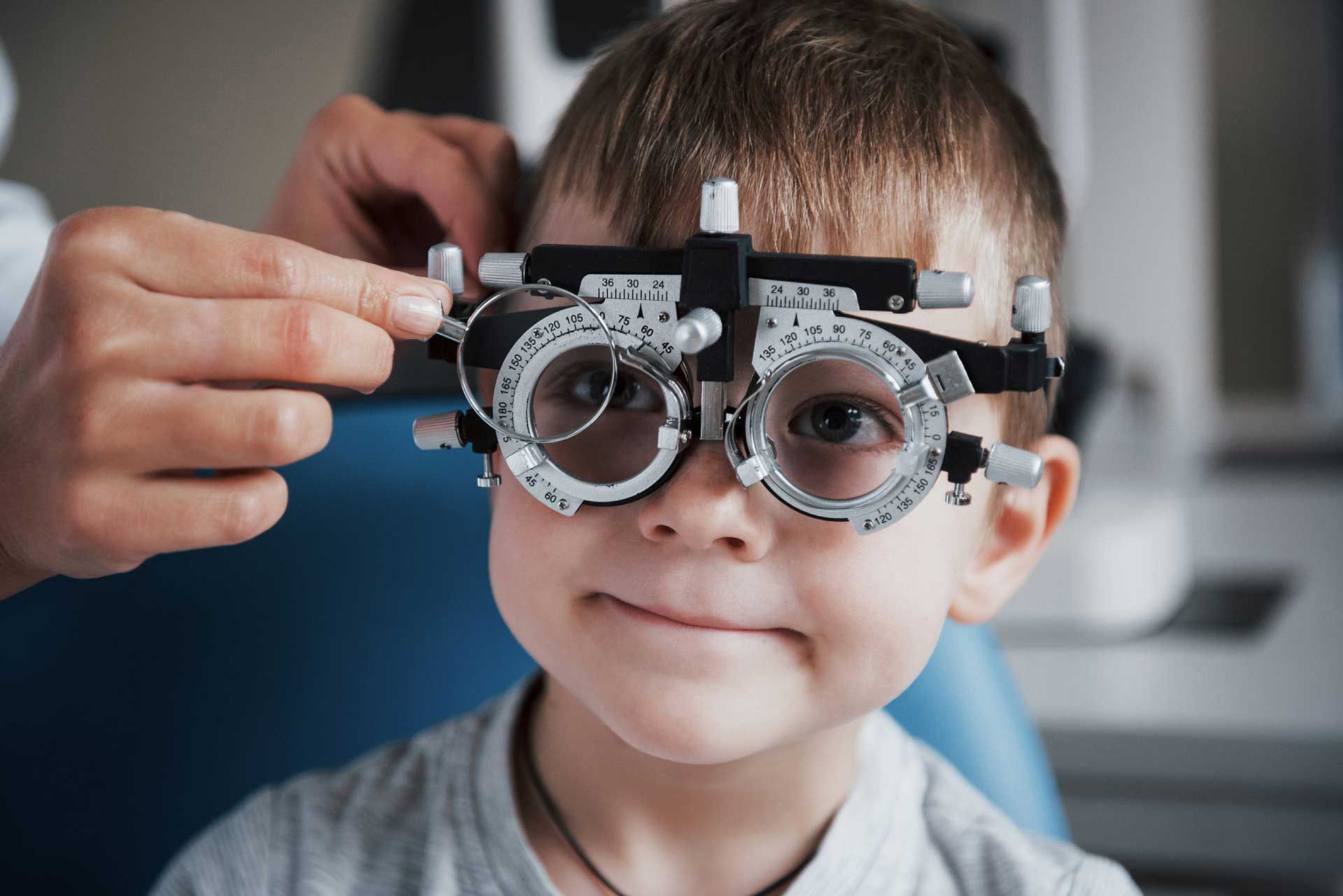 Young boy wearing a phoropter at an eye exam, a doctor adjusting the lenses, smiling.